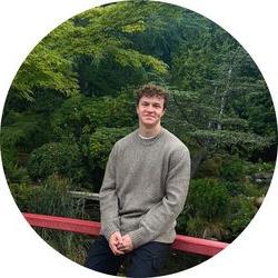 Tutor Josh wearing a sweater and leaning on a red metal railing in a park with trees and a pond in the background.